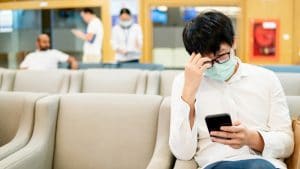 Male Asian patient wearing surgical mask using smartphone at waiting area in hospital or medical center. Medical exam or body check up. Wuhan coronavirus outbreak prevention. Health care concept
